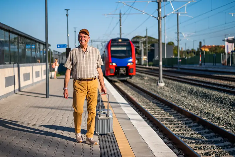 Älterer Mann mit Rollkoffer geht am Bahnsteig entlang, dahinter fährt ein Zug ein