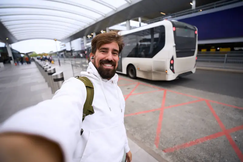 Lächelnder Mann mit Rucksack macht ein Selfie vor einem Bus am Flughafen