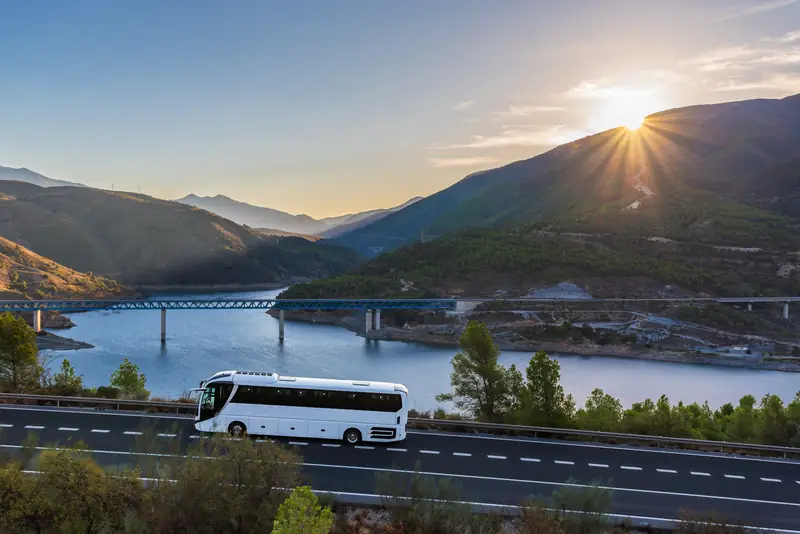 Reisebus fährt bei Sonnenuntergang auf einer Straße an einem Stausee